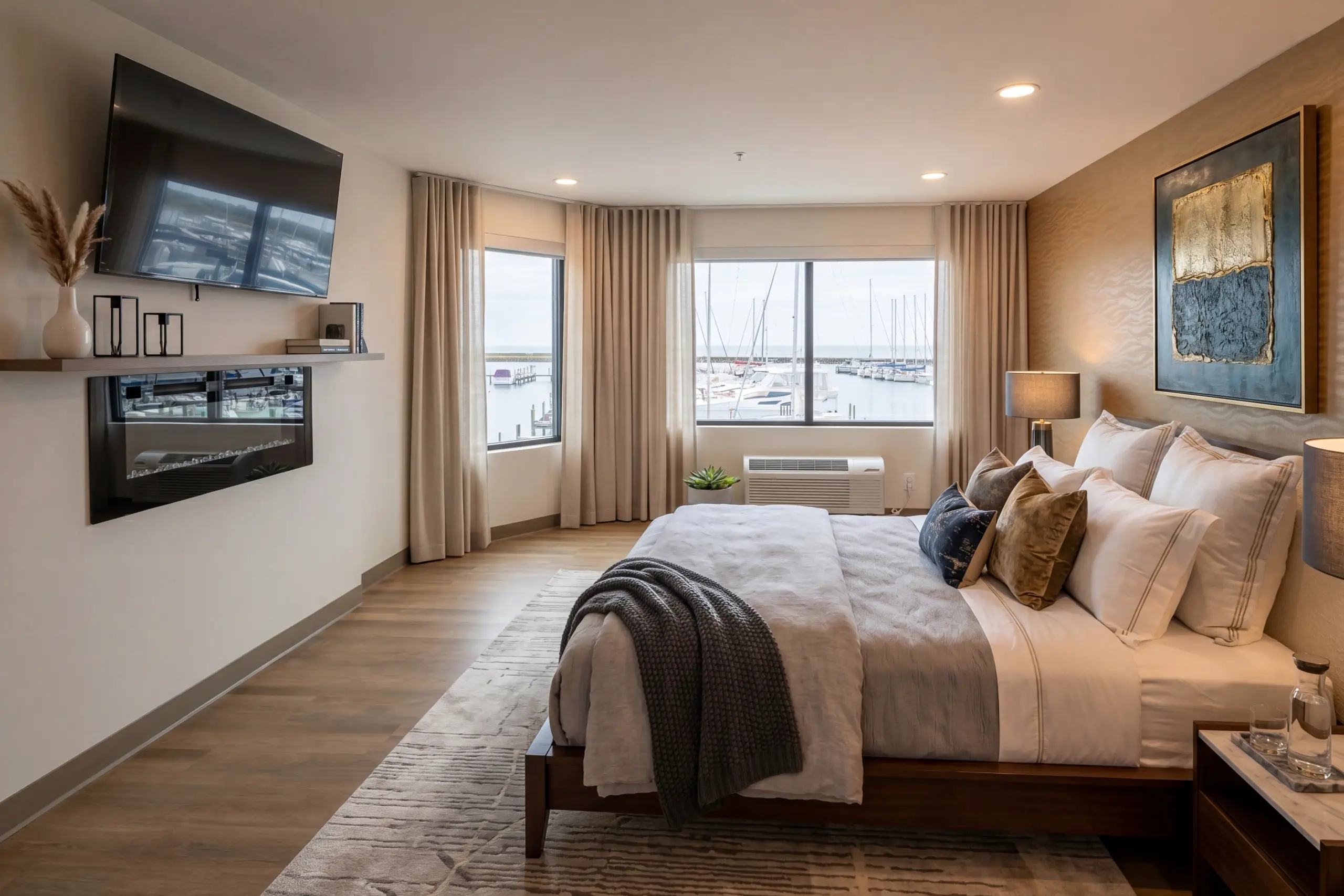 A contemporary hotel room with a large bed, featuring views of a bustling marina through expansive windows. A TV and electric fireplace are mounted on the adjacent wall.