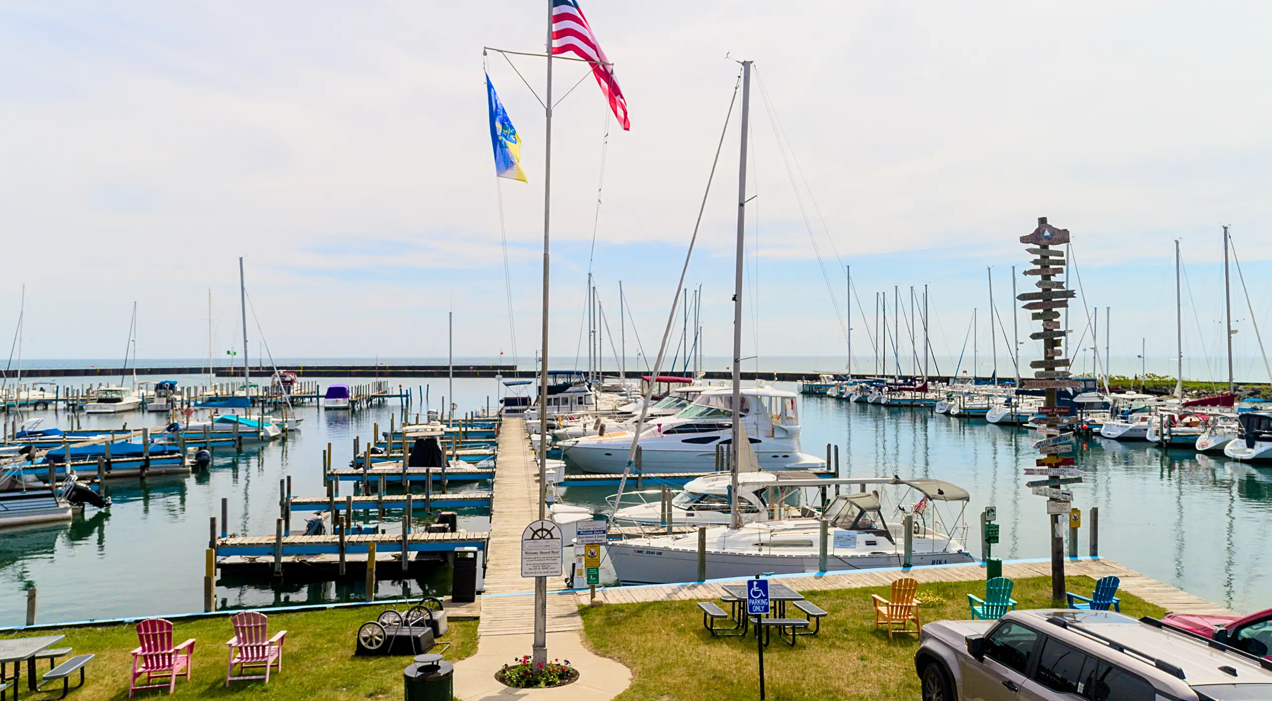 A wide view of a sunny marina filled with docked sailboats and motorboats, featuring a wooden pier, a directional signpost, and colorful chairs on the shore.