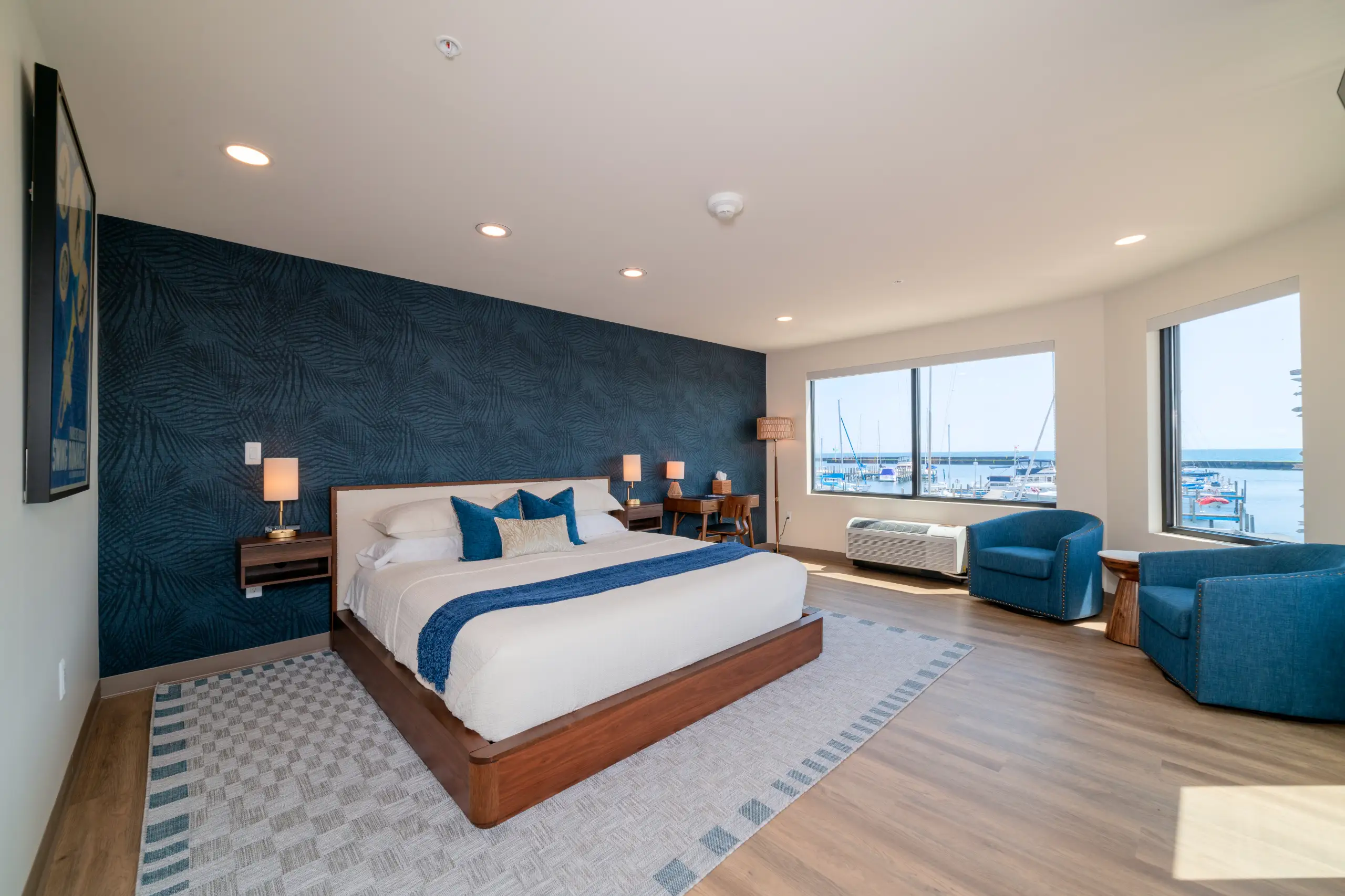 A king hotel room with blue accents, dark blue leaf wallpaper, and a large window providing a panoramic view of boats docked in a harbor.