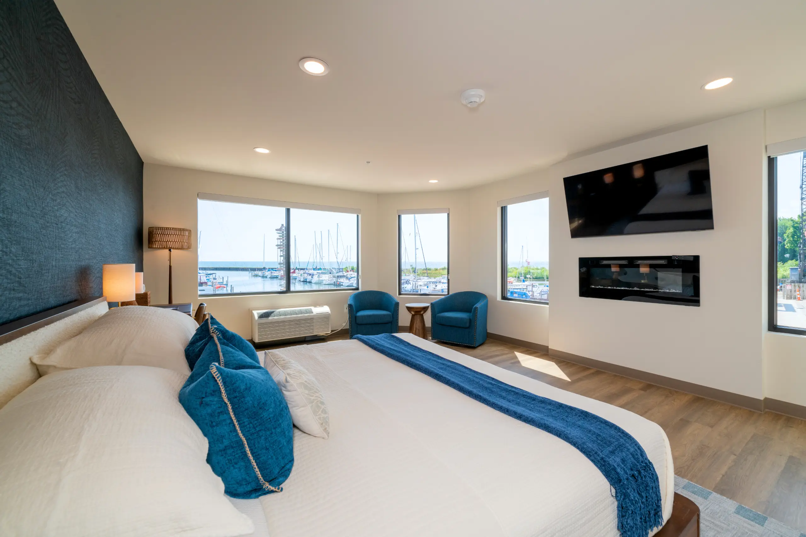 A modern king hotel room features blue accents, a wall-mounted TV, and a fireplace. The expansive windows offer a bright, scenic view of a harbor filled with docked sailboats.