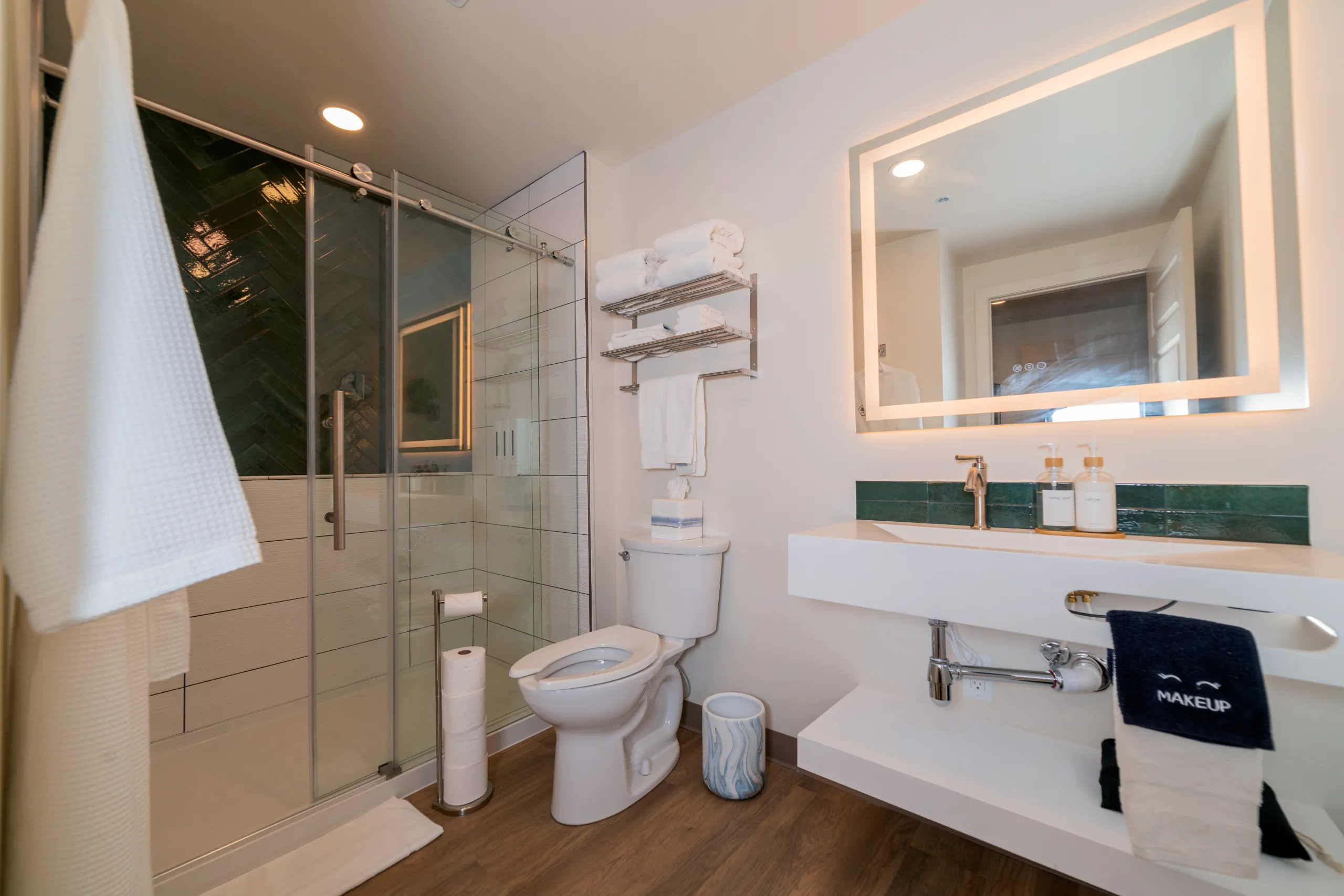 A clean, modern hotel bathroom with a glass walk-in shower, dark green tiled wall, white toilet, and a sleek floating sink under an illuminated mirror.
